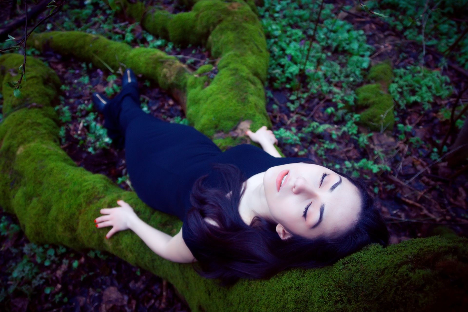 Fashion shot of a dark-haired beautiful woman lying on a moss-covered big tree roots.
The young woman is a very beautiful and attractive. The girl is dressed in black dress. She is carefree and serene